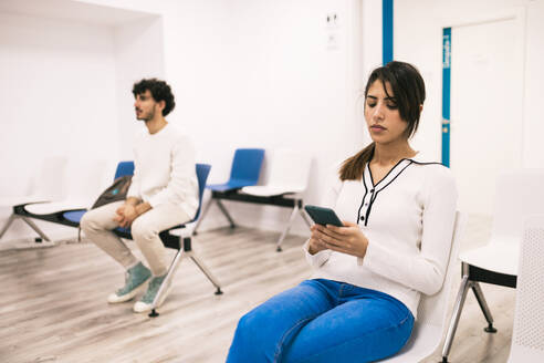 A young woman focused on her smartphone in a waiting room, with another person seated in the background. - ADSF53683