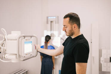 A male patient stands next to a radiographic machine in a clinic while a technician prepares the equipment in the background. - ADSF53691