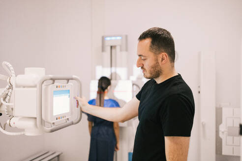A male patient stands next to a radiographic machine in a clinic while a technician prepares the equipment in the background. - ADSF53691