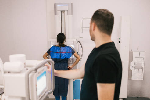 A patient wearing a protective lead apron prepares for an x-ray procedure as the technician adjusts the machine. - ADSF53694