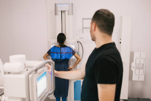 Ein Patient, der eine Bleischürze trägt, bereitet sich auf eine Röntgenuntersuchung vor, während der Techniker das Gerät einstellt. - ADSF53694