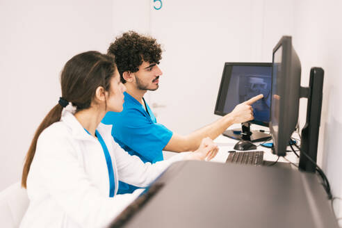Two healthcare workers in blue scrubs are focused on a computer screen, discussing medical data in a bright office setting. - ADSF53699