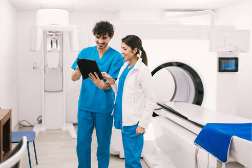 Two medical staff members, a man and a woman, are engaged in a discussion over a digital tablet in a well-equipped radiology room with an MRI scanner in the background. - ADSF53701