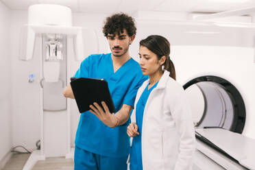 Two healthcare workers, a male in scrubs and a female in a lab coat, are intently reviewing a digital tablet in a modern hospital setting, possibly near MRI equipment. - ADSF53703