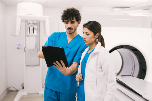 Two healthcare workers, a male in scrubs and a female in a lab coat, are intently reviewing a digital tablet in a modern hospital setting, possibly near MRI equipment. - ADSF53703