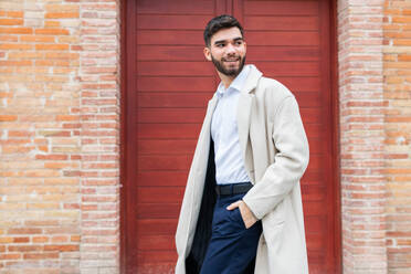 A stylish young man in business attire smiles confidently while standing against a red door backdrop, exuding a sense of casual professionalism in his beige coat and navy trousers - ADSF53707