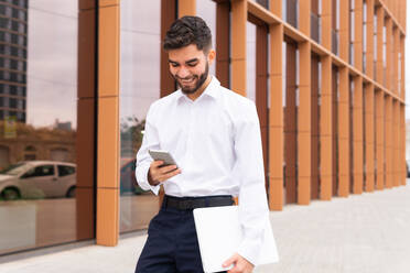 A cheerful businessman in a white shirt checks his phone while holding a laptop, standing against a modern architectural backdrop - ADSF53708