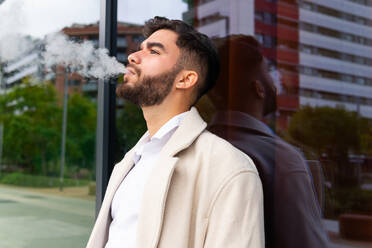 A contemplative businessman exhales smoke while standing thoughtfully near a reflective glass building, dressed in a stylish beige coat - ADSF53710