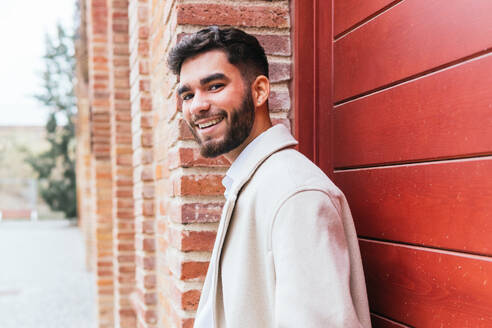 Charming businessman in a stylish coat smiling and leaning against a red brick wall, radiating confidence and ease - ADSF53712