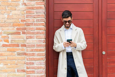 A joyful businessman in a beige coat and sunglasses using his phone, leaning against a brick wall next to a red door - ADSF53714