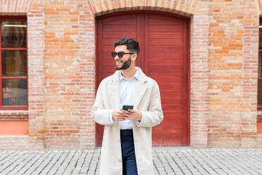 Cheerful businessman in a trench coat and sunglasses browsing on his phone, standing in front of a classic brick building with a red door - ADSF53715
