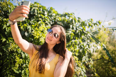 A cheerful young woman in a sunny garden takes a selfie with a cellphone capturing the moment with joy - ADSF53716