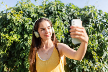 A cheerful young lady in a yellow tank top takes a selfie with her smartphone while wearing white over-ear headphones, standing before lush green foliage - ADSF53717