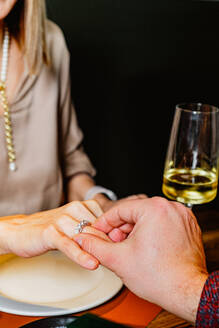 A close-up of a man's hand placing an engagement ring on a woman's finger during a romantic dinner, with a wine glass in the background - ADSF53729