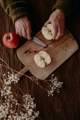 A close-up view of aged hands cutting a fresh apple on a rustic wooden cutting board, with a serene and homely atmosphere - ADSF53743