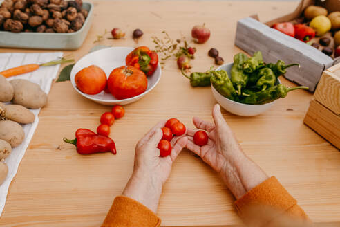 Hands sorting cherry tomatoes with assorted fresh vegetables on a wooden table, prepping for a healthy meal - ADSF53744