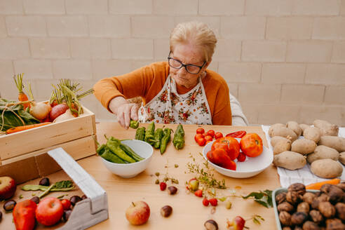 A senior woman with glasses meticulously chops vegetables in a well-lit kitchen, surrounded by an array of healthy produce - ADSF53745