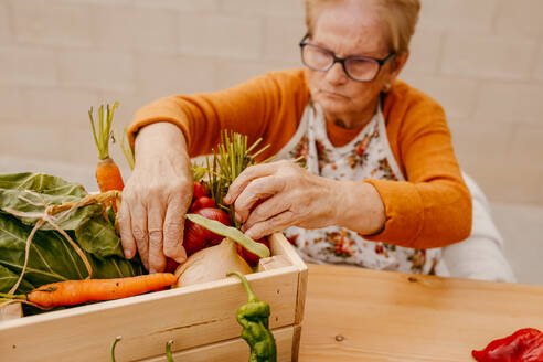 Elderly person organizing a variety of freshly harvested vegetables in a wooden box on a tabletop - ADSF53748