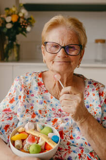 A cheerful elderly lady with glasses smiles as she holds a pink lollipop and a bowl of colorful candies - ADSF53757