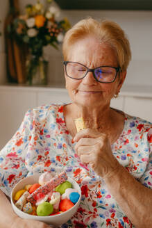 A content elderly lady holds a bowl of vibrant candies while eating a biscuit, showcasing delightful small pleasures at an advanced age - ADSF53760