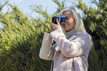 A smiling woman in her 50s enjoying photography with a vintage camera in a natural setting - ADSF53761