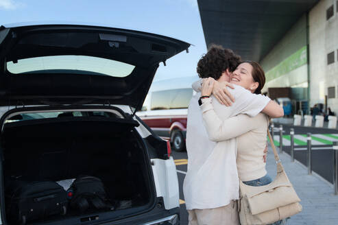 A man and woman embrace warmly in front of a car with the trunk open at the airport, conveying a heartfelt farewell. - ADSF53769