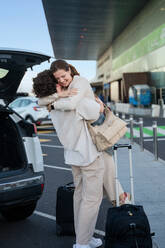 Woman eagerly embraces a traveler by the car at the airport, expressing joy and affection, with luggage beside them. - ADSF53771