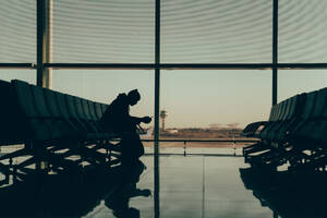 A back-view silhouette of an unrecognizable man sitting alone, checking his phone, waiting for his flight at an airport terminal with a view of the control tower in the background. - ADSF53775