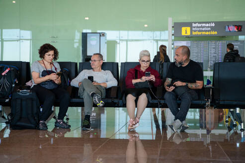 Two men and two women engrossed in their smartphones while waiting in an airport lounge, reflecting modern travel habits. - ADSF53778