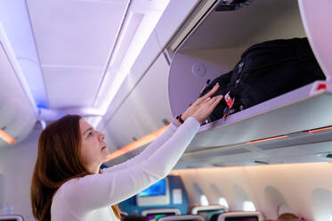 Side view of woman places her bag in the overhead storage on a flight to Malaysia, beginning her travel adventure. - ADSF53782