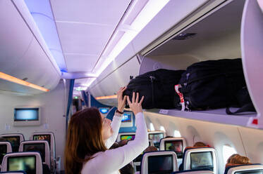 A woman is placing her bag into the overhead storage compartment on an airplane during a flight, possibly heading to Malaysia. - ADSF53784