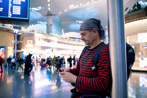 A man waits in the bustling terminal of an airport, engrossed in his phone while checking his flight information, clearly embodying the modern travel experience. - ADSF53787