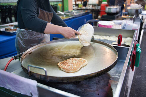 A cropped unrecognizable street vendor in Malaysia skillfully prepares Roti Canai, a popular Malaysian flatbread, on a large griddle at an outdoor market. - ADSF53793