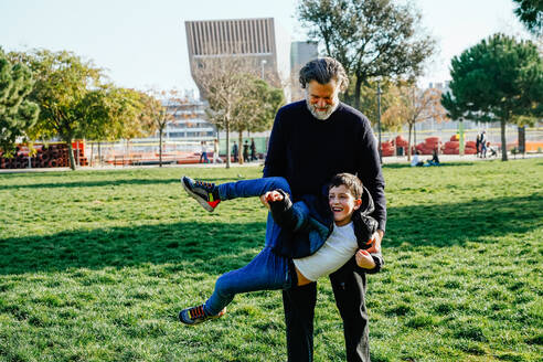 A happy child enjoys a playful moment with their grandfather, spinning around in a sunny park setting - ADSF53809