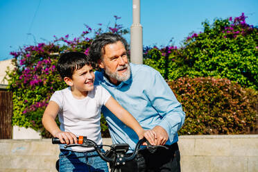 An elderly man smiling as he supports a young boy learning to ride a bicycle in a sunny outdoor setting - ADSF53813