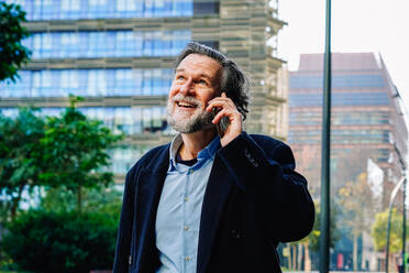 Mature businessman enjoys a conversation on his mobile phone, with office buildings in the backdrop, exhibiting the fusion of technology and everyday life - ADSF53816
