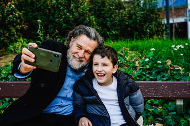 A senior man with a grey beard and a young boy smile joyfully while taking a selfie together on a wooden park bench surrounded by greenery - ADSF53819