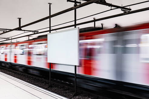 A blurred red train speeds past an empty advertising billboard at a modern train station, hinting at the pace of urban life - ADSF53821