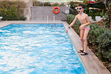 A cheerful young boy wearing goggles and swim shorts stands ready to dive into a shimmering outdoor swimming pool, with a lifebuoy nearby - ADSF53822