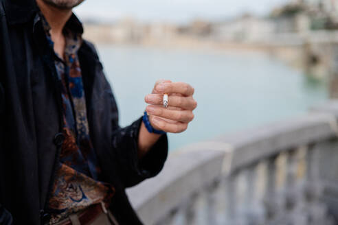 Close-up of a man's hand presenting a stylish ring against a blurred seaside backdrop, capturing a moment of elegance and fashion - ADSF53825