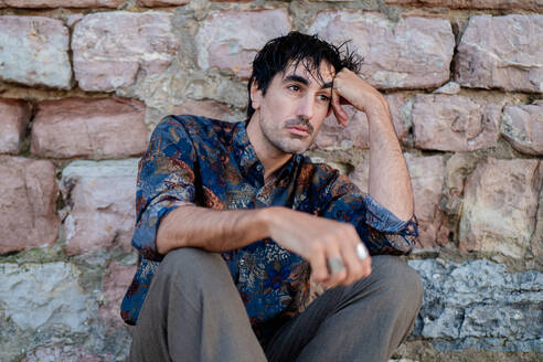 Contemplative young man sitting on rocky ground against a stone wall - ADSF53827