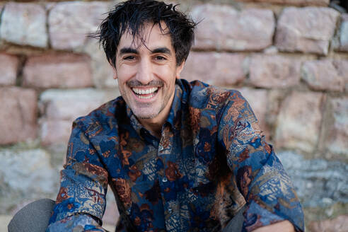 Cheerful man with a genuine smile, wearing a patterned shirt and wet hair, sitting against a stone wall backdrop - ADSF53831