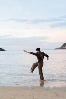 Man balancing playfully on one leg at the beach with outstretched arms during dusk - ADSF53833