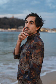 A pensive man stands on the shoreline, smoking a cigarette as evening falls, with soft focus lights in the background - ADSF53834