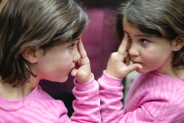 A young girl in a pink top contemplates her reflection in a mirror, displaying curiosity and innocence. - ADSF53836