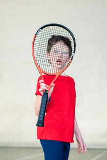 A young boy poses with a tennis racket, half obscured, ready for a game on a sporty weekend. - ADSF53837