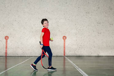 A young boy smiles as he holds a tennis racquet and ball, ready for a match on an indoor court. - ADSF53838