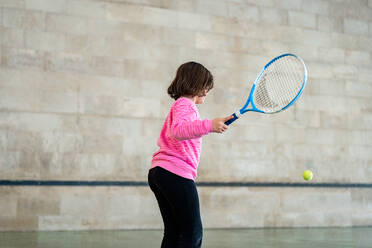 Back view of unrecognizable focused young girl practices her tennis swing with a racket, aiming at a yellow ball in an indoor setting. - ADSF53839