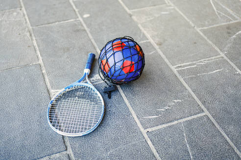 A badminton racket lying next to a soccer ball in a blue and red net on a concrete surface, symbolizing outdoor activities. - ADSF53841