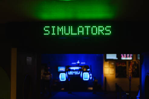 Grünes LED-Schild mit der Aufschrift SIMULATORS in einem schwach beleuchteten Spielhallenraum mit blauen Neonröhren im Hintergrund - ADSF53842